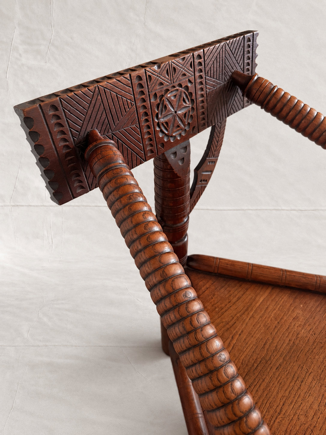 Close-up of a carved wooden chair back and armrests, showing detailed geometric patterns and ribbed turned wood on a light neutral background.