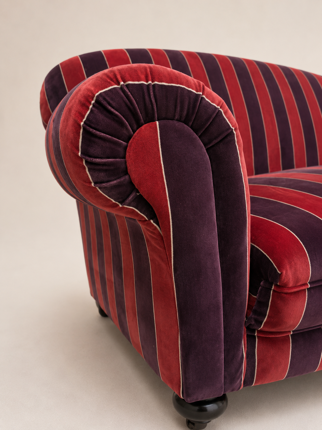 Close-up of a classic Chesterfield armchair upholstered in red and dark purple striped velvet fabric with thin white piping, on a light beige background.