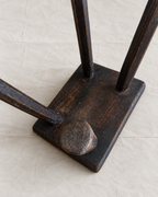 Close-up of a dark stained wooden stand or rack base with two vertical posts and a round peg-like support, photographed on a light beige textured surface.