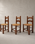Three rustic wooden ladder chairs with woven straw seats, arranged side-by-side against a textured light gray backdrop.