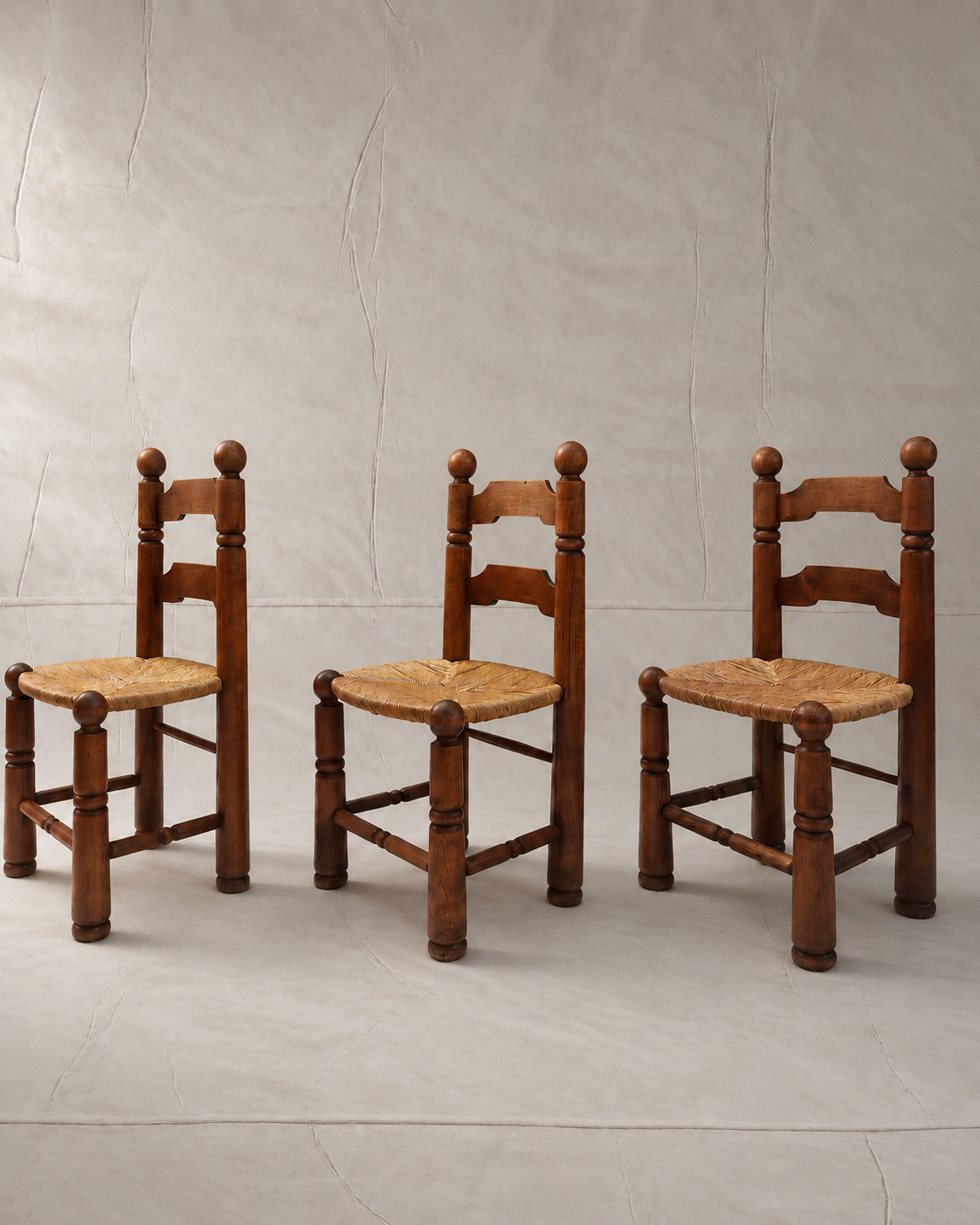 Three rustic wooden ladder chairs with woven straw seats, arranged side-by-side against a textured light gray backdrop.