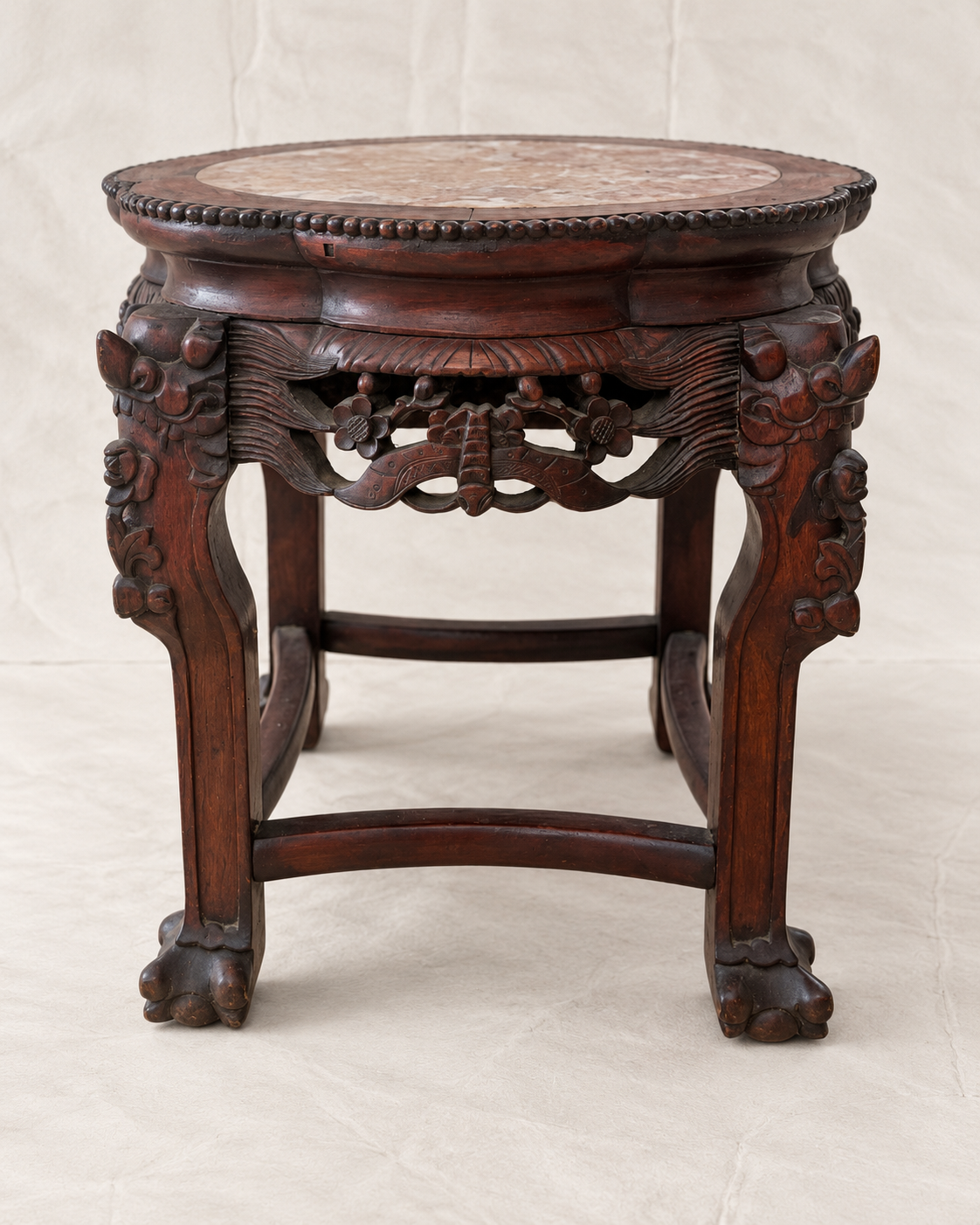 An ornate dark brown carved wooden stool with a round stone-topped surface and decorative scrollwork details, photographed against a light beige fabric backdrop.