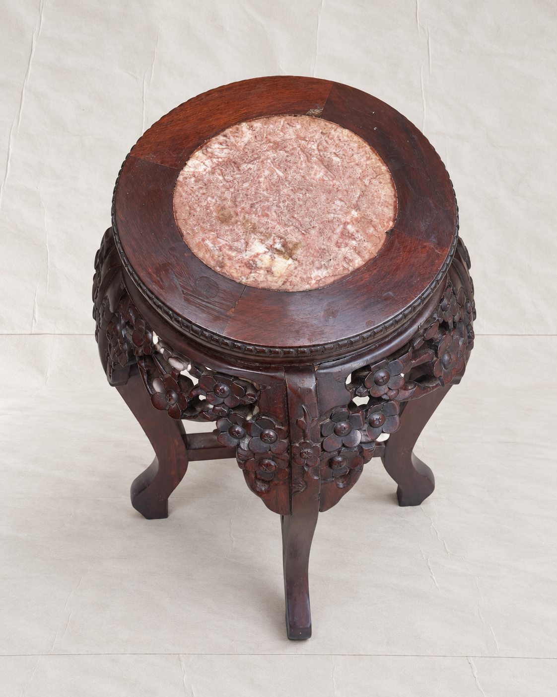 A round wooden stool with a carved dark reddish-brown floral base and a circular pink stone inlay on the top, photographed against a light beige fabric backdrop.