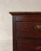 Close-up of a dark brown wooden chest of drawers showing the top corner, drawer front, and brass hardware against a light textured backdrop.