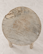 A rustic round wooden stool with a distressed, weathered finish, featuring a thick circular seat and two turned legs, photographed from a top-down angle on a textured off-white background.