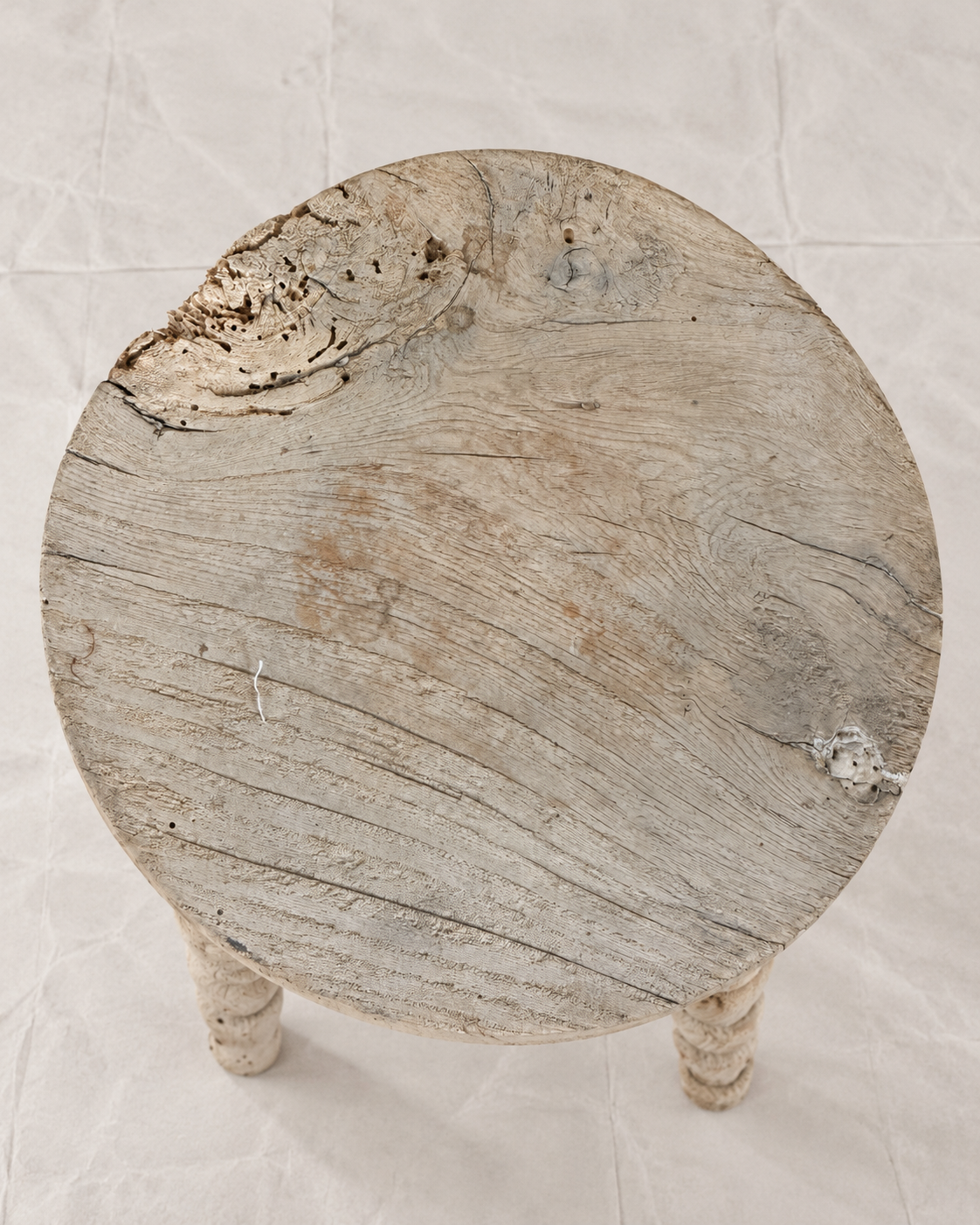 A rustic round wooden stool with a distressed, weathered finish, featuring a thick circular seat and two turned legs, photographed from a top-down angle on a textured off-white background.