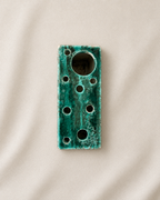 A rectangular ceramic bud vase with a distressed teal-green glaze and multiple circular cutouts, photographed on a neutral beige fabric background.