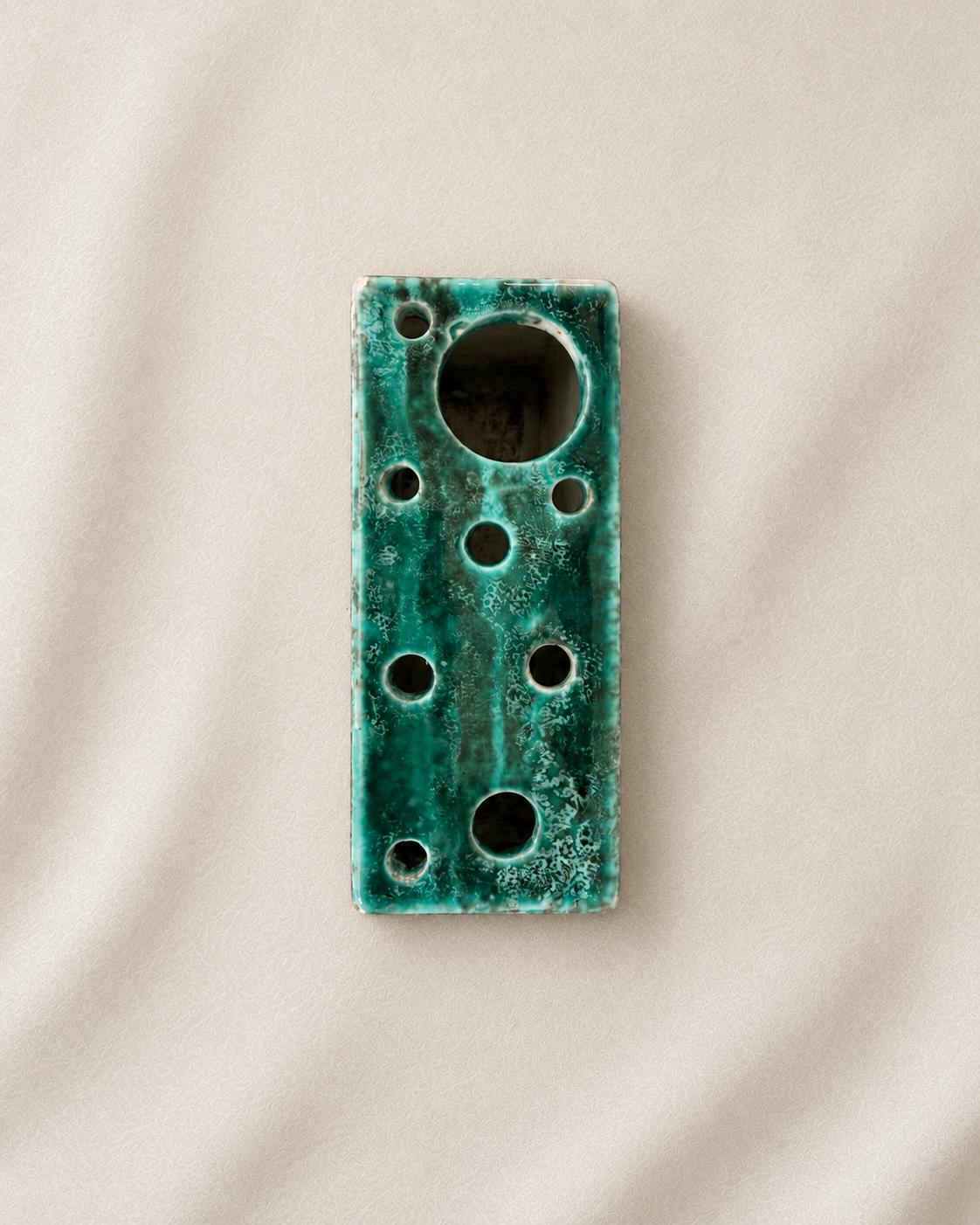 A rectangular ceramic bud vase with a distressed teal-green glaze and multiple circular cutouts, photographed on a neutral beige fabric background.