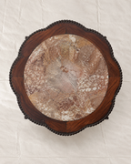 An overhead view of a round wooden table top featuring a natural brown and beige marble inlay surrounded by a dark reddish-brown wood frame with a scalloped edge and decorative bead-like trim.