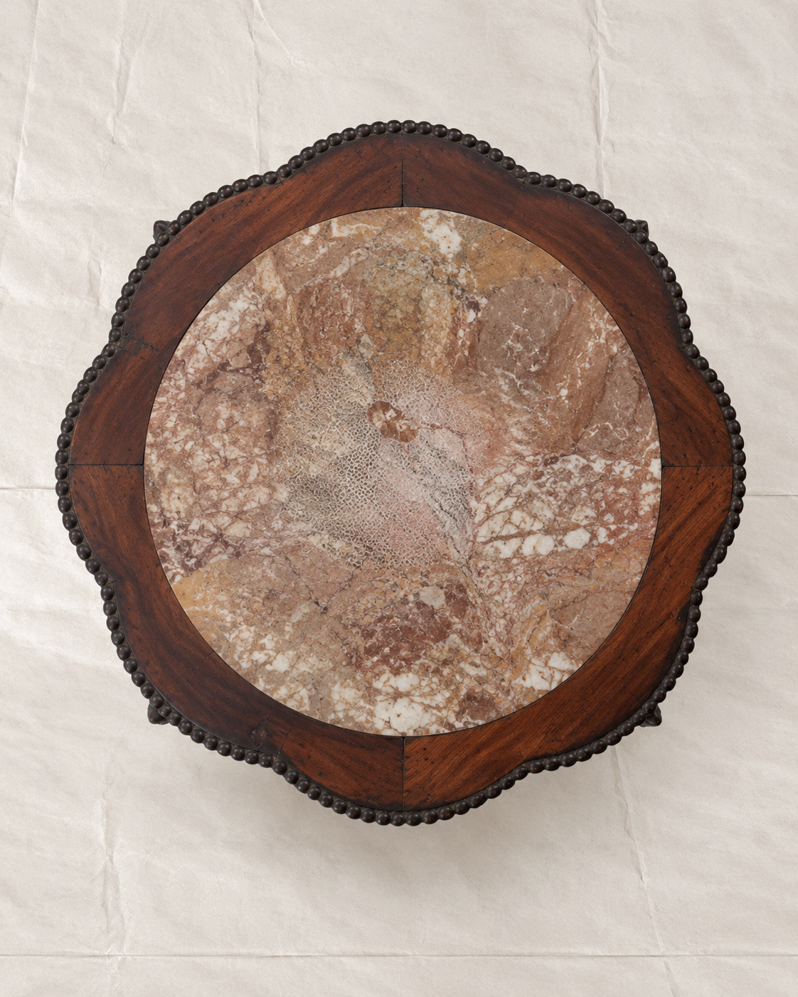 An overhead view of a round wooden table top featuring a natural brown and beige marble inlay surrounded by a dark reddish-brown wood frame with a scalloped edge and decorative bead-like trim.