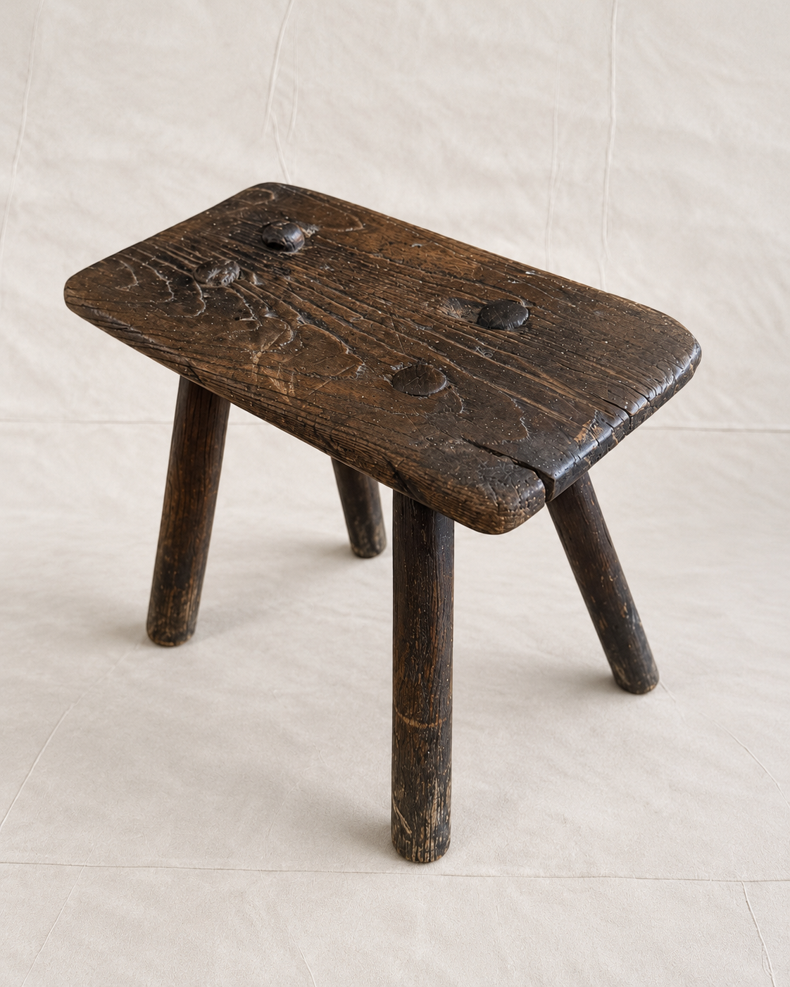 A rustic dark brown wooden stool with a thick rectangular seat and three legs, photographed against a neutral off-white fabric backdrop.