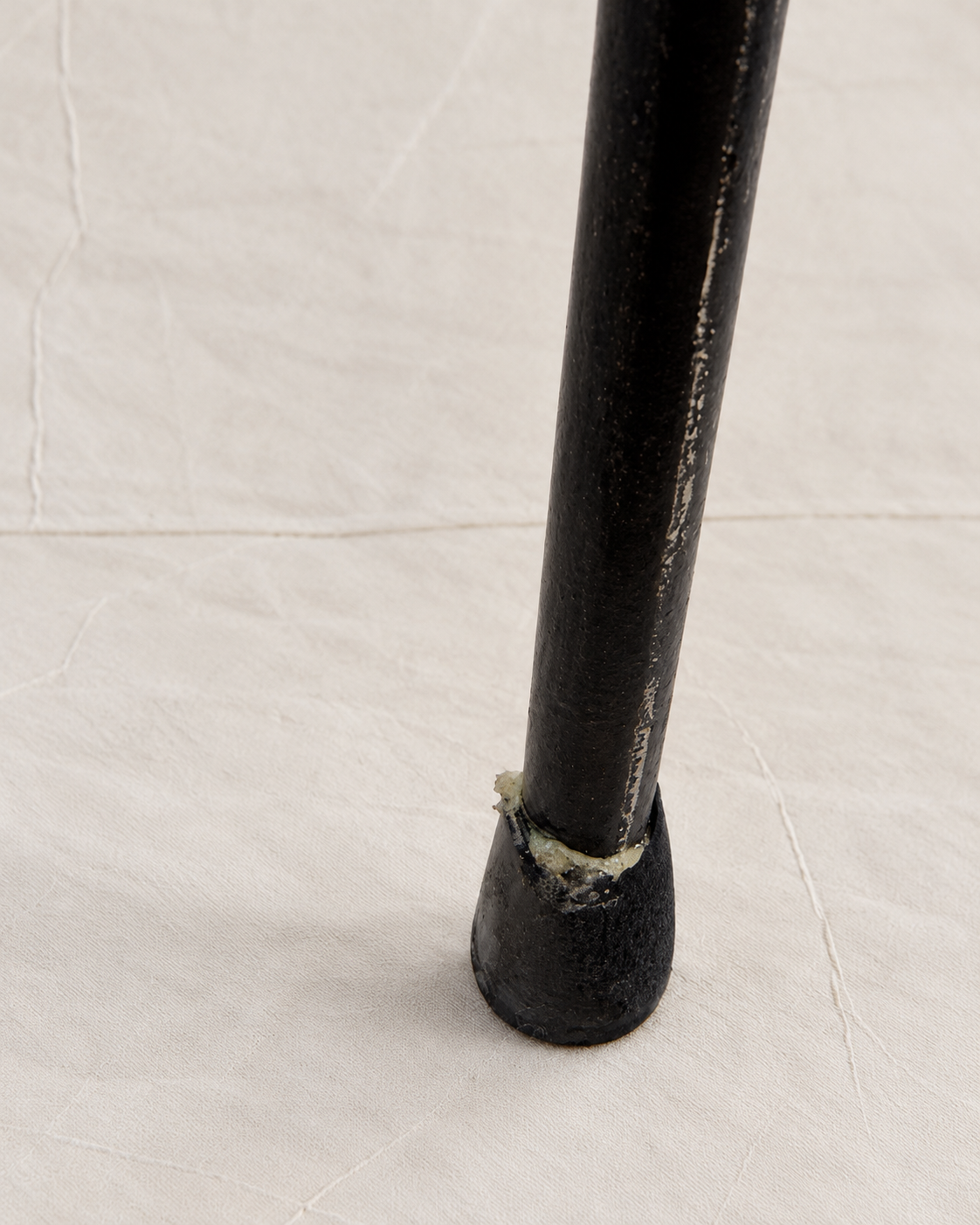 A close-up of a black metal table leg with a worn, chipped section near the base, set against a textured off-white background.