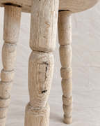Close-up of a distressed, whitewashed wooden stool leg with a turned spindle design, showing the rustic wood grain and chipped paint details against a neutral light background.
