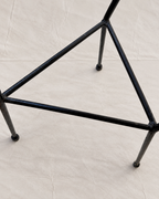 A close-up of a black metal triangular stand or frame with four legs, photographed on a light beige fabric surface.