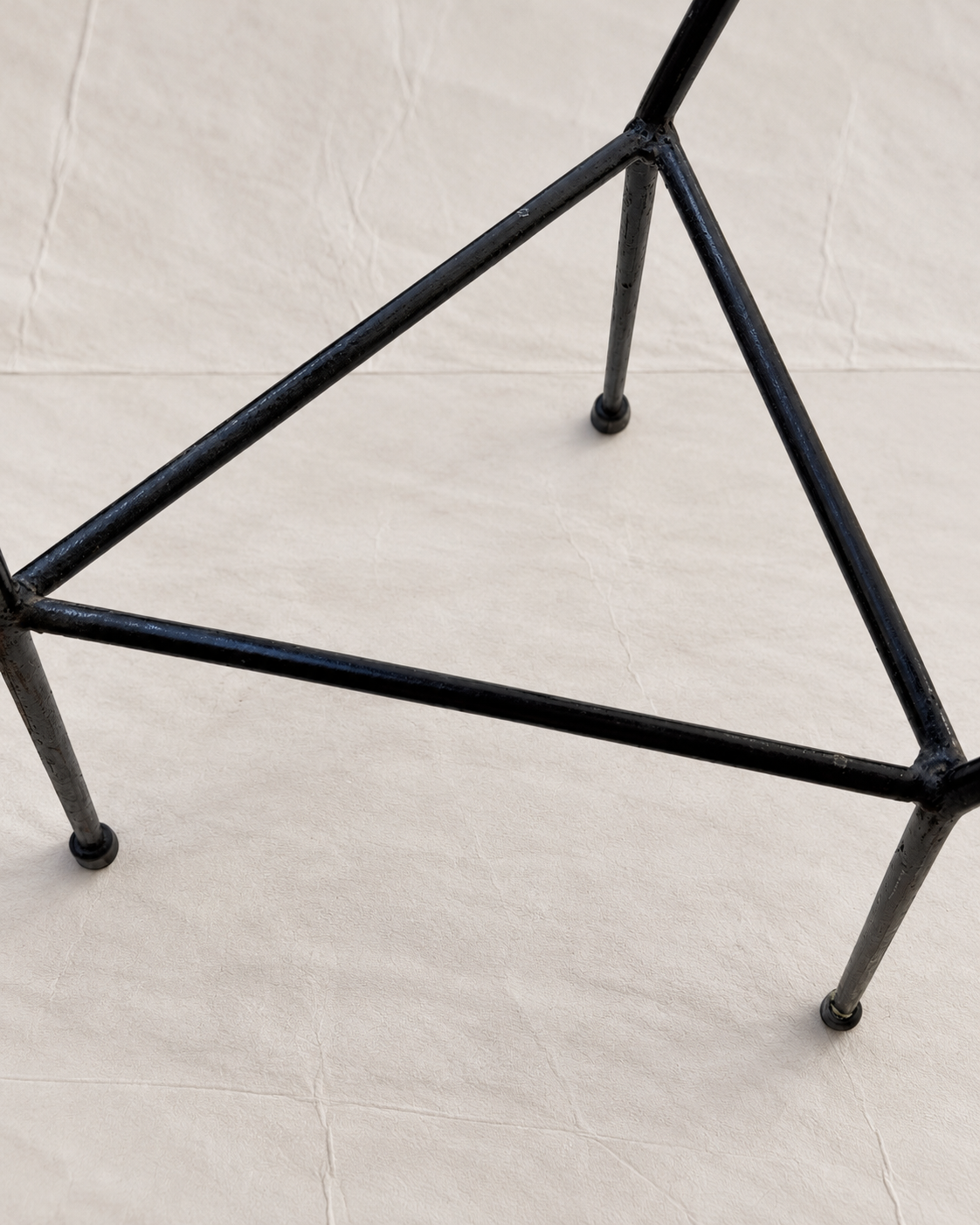 A close-up of a black metal triangular stand or frame with four legs, photographed on a light beige fabric surface.