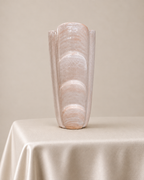 A tall, sculptural ceramic vase with a glossy, textured finish in a warm cream-beige color, displayed on a draped satin tablecloth against a neutral beige background.