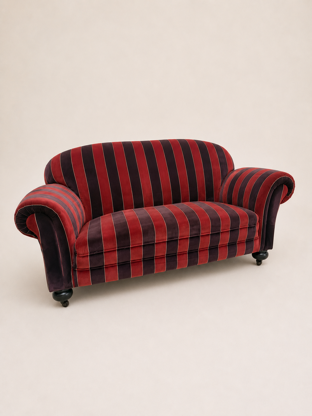 A vintage loveseat sofa with red and black striped velvet upholstery, featuring rolled arms, dark wooden legs, and small black caster wheels, photographed against a plain beige background.