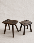 Two rustic dark brown wooden stools with a weathered finish, displayed on a light beige textured background.