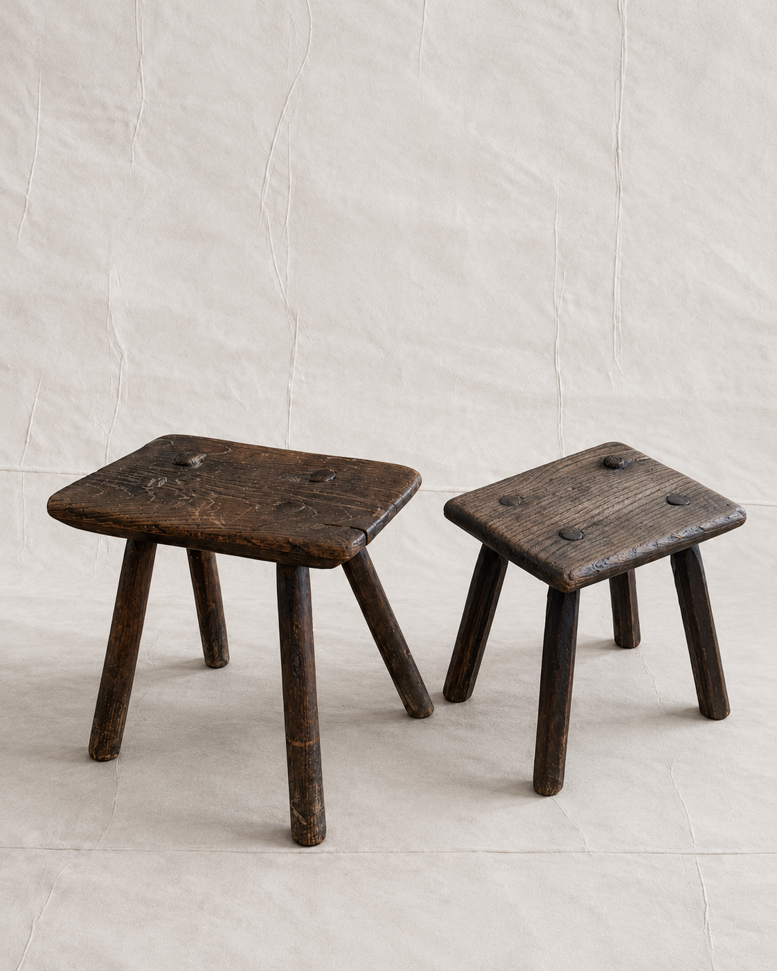 Two rustic dark brown wooden stools with a weathered finish, displayed on a light beige textured background.