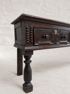 An antique dark wood chest or side table with a turned leg, decorative drawer fronts, and brass hardware, photographed against a neutral draped fabric backdrop.