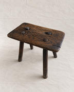 A rustic dark brown wooden stool with a thick square seat and four legs, featuring four metal pegs on top, set against a light beige fabric backdrop.