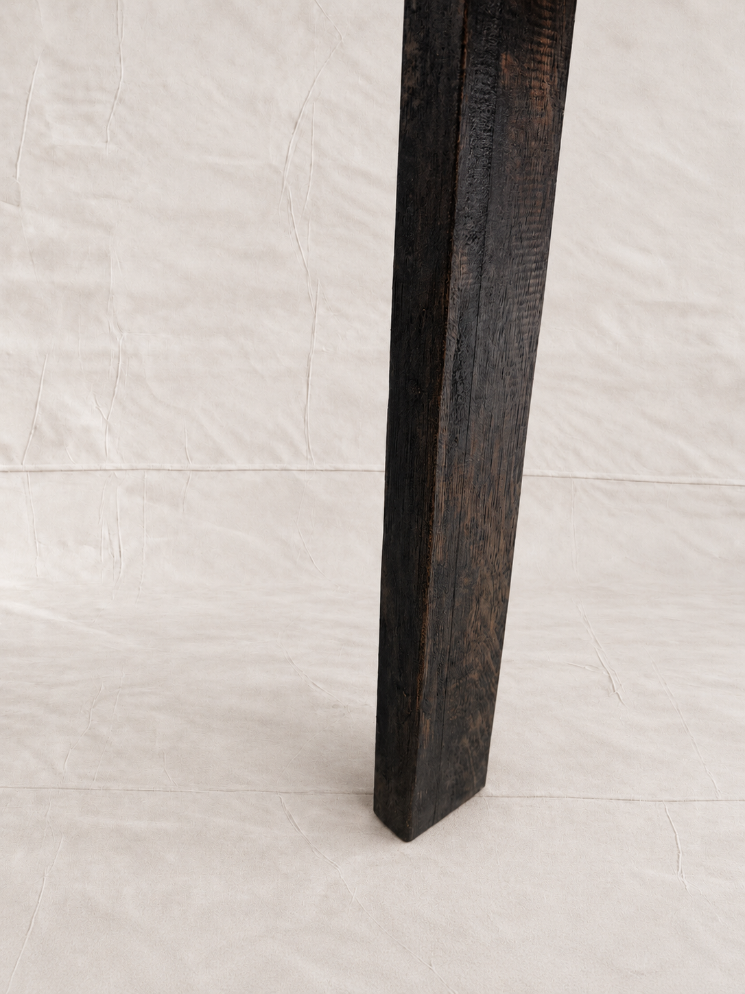 A close-up of a dark stained wooden table leg standing vertically against a light beige fabric backdrop.