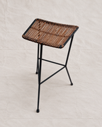 A modern bar stool with a square woven brown seat and a black metal frame, photographed against a neutral beige fabric backdrop.