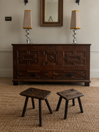 A dark carved wooden chest of drawers in a styled room, flanked by two small wooden stools on a woven rug, with two table lamps and a mirror above.