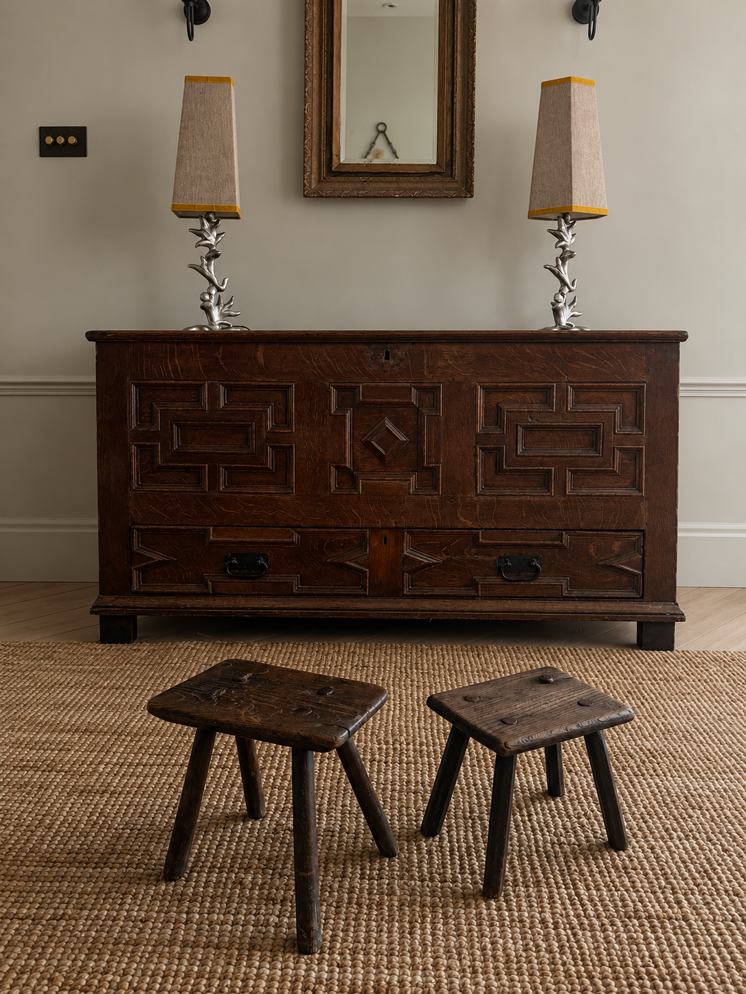 A dark carved wooden chest of drawers in a styled room, flanked by two small wooden stools on a woven rug, with two table lamps and a mirror above.