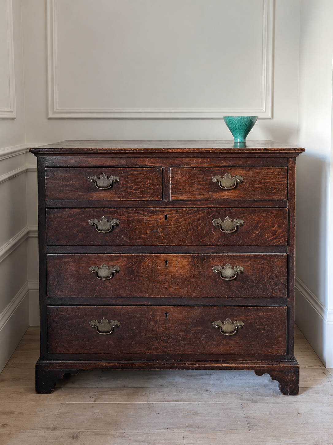 A dark brown wooden chest of drawers with five drawers and ornate metal handles, set against a white paneled wall on a light wood floor, with a small green vase on top.