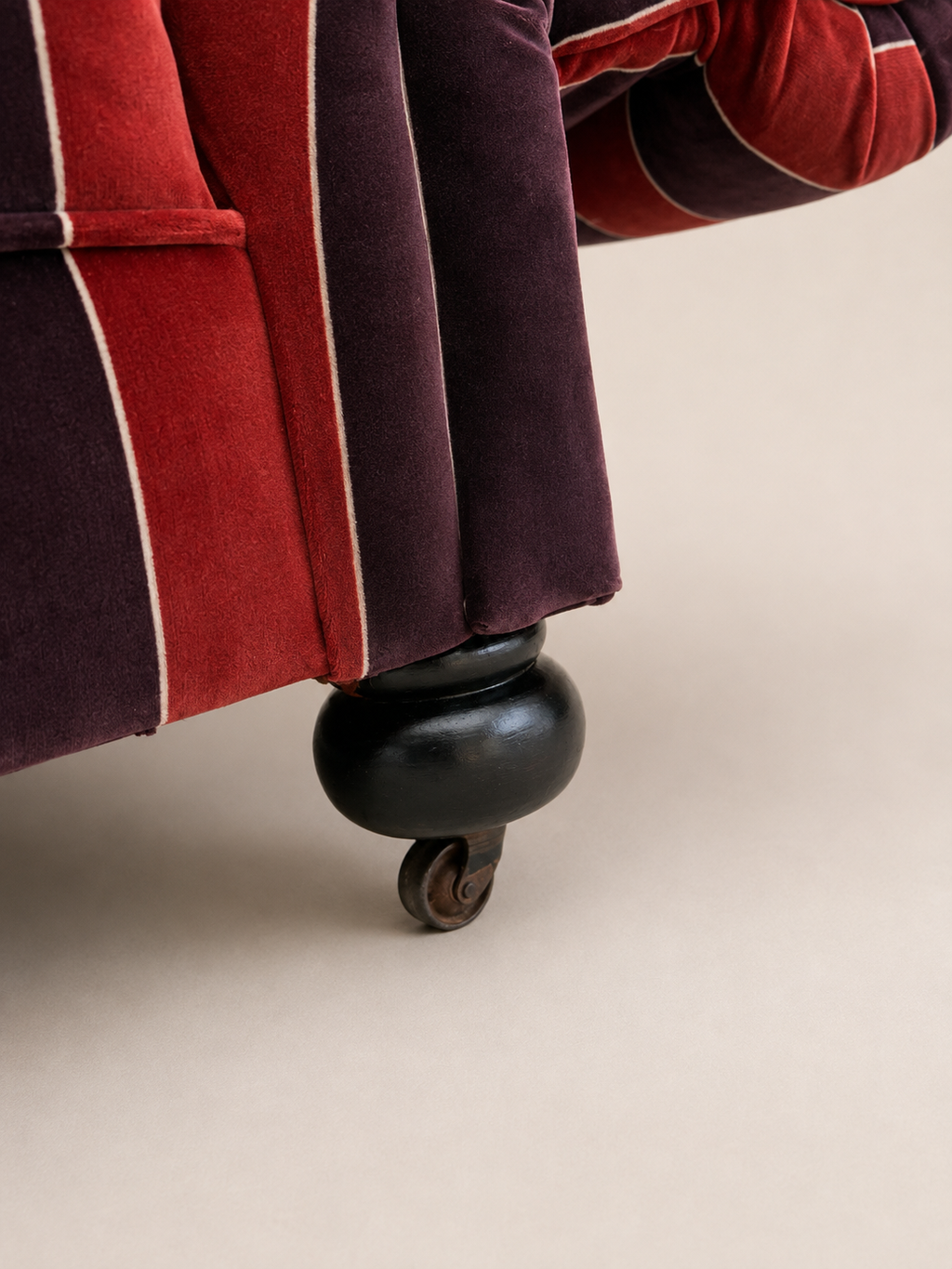 Close-up of a velvet upholstered armchair or sofa leg, showing deep red and dark plum vertical stripes with thin cream piping, a black turned wooden footstool, and a small wheel caster on a warm beige background.