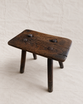A rustic dark brown wooden stool with a rectangular seat, three legs, and three metal pegs on top, set against a neutral beige fabric backdrop.