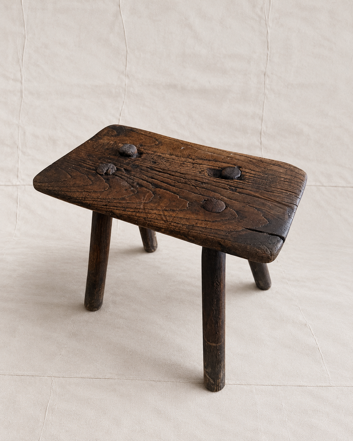 A rustic dark brown wooden stool with a rectangular seat, three legs, and three metal pegs on top, set against a neutral beige fabric backdrop.