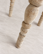 A close-up of a distressed, light beige wooden table leg with carved details, standing on a light beige tiled floor.