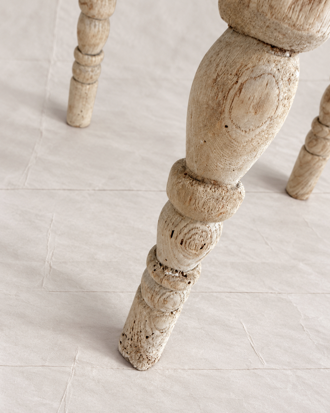 A close-up of a distressed, light beige wooden table leg with carved details, standing on a light beige tiled floor.