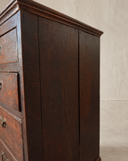Close-up of a dark brown wooden chest of drawers showing the side panel, drawer front, and brass hardware against a light beige wall.