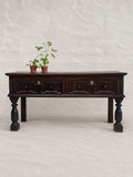 A dark brown wooden console table with carved drawer fronts and turned legs, styled with two small potted plants on top, set against a neutral off-white fabric backdrop.
