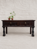 A dark brown wooden console table with carved drawer fronts and turned legs, styled with two small potted plants on top, set against a neutral off-white fabric backdrop.