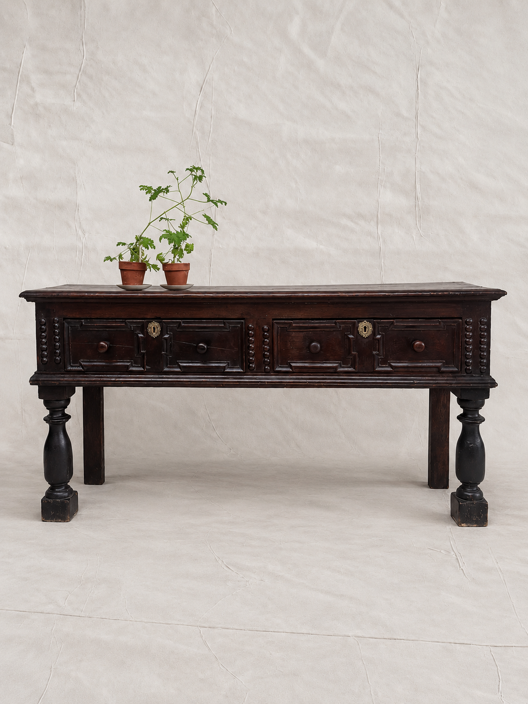 A dark brown wooden console table with carved drawer fronts and turned legs, styled with two small potted plants on top, set against a neutral off-white fabric backdrop.