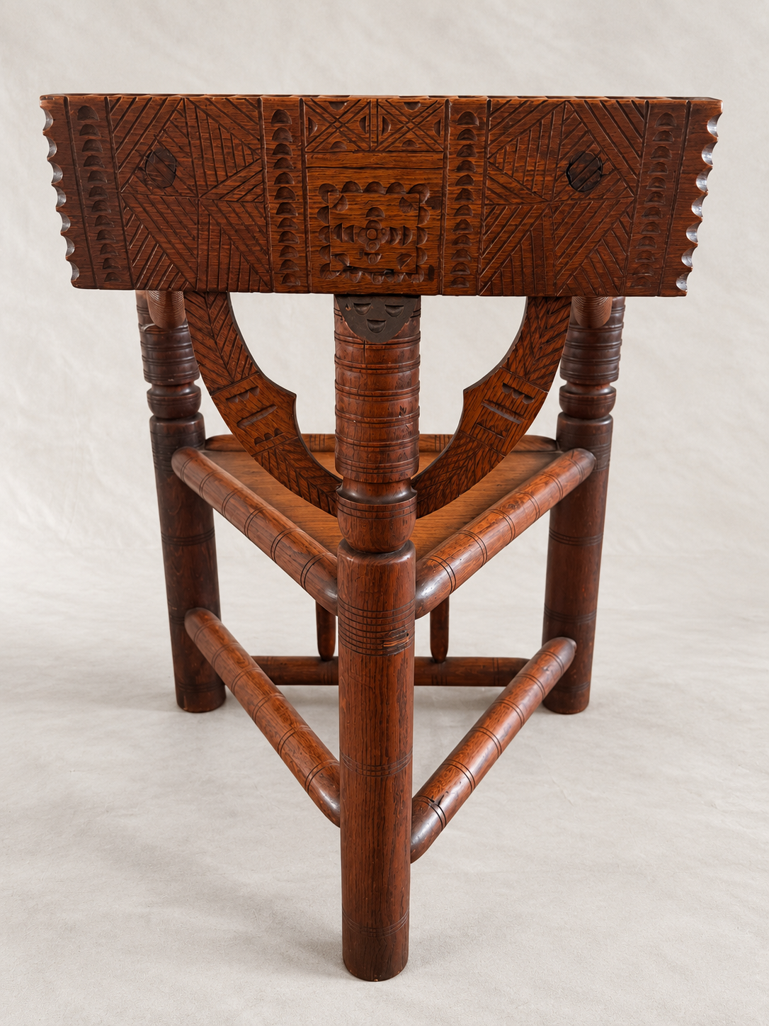 A dark brown carved wooden stool with geometric patterns on the seat and backrest, featuring turned legs and a woven seat panel, photographed against a neutral light gray background.