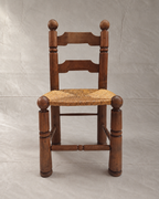 A rustic Charles Dudouyt wooden ladder-back chair with a woven rush or cane seat, photographed against a neutral gray fabric backdrop.