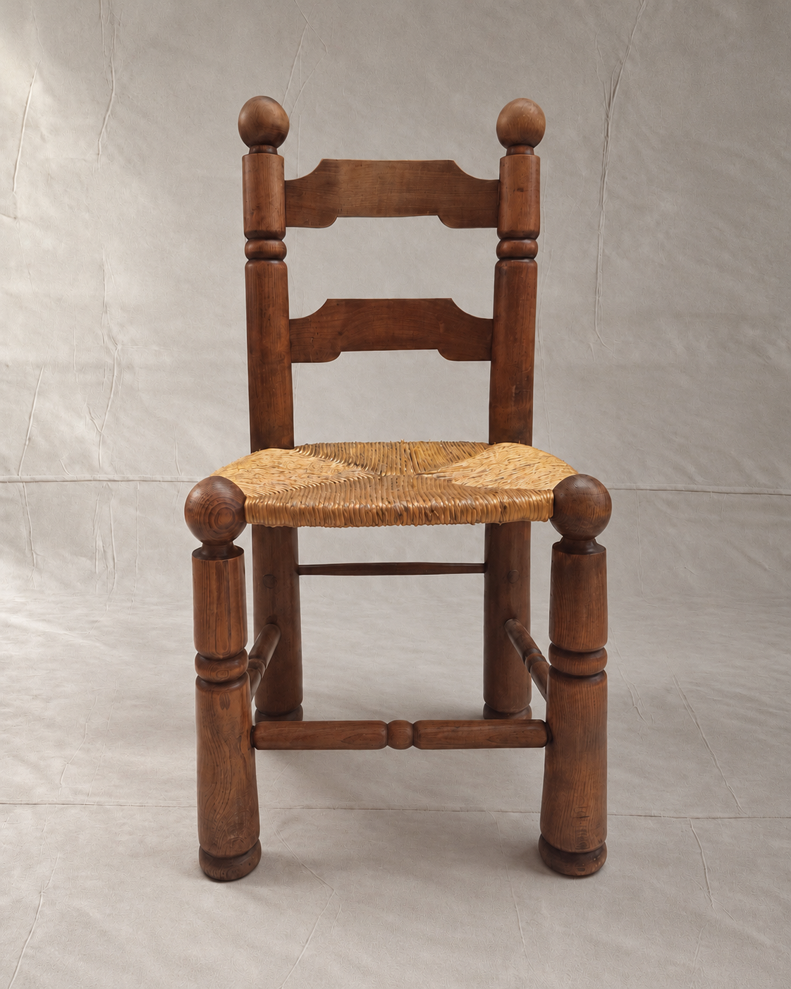 A rustic Charles Dudouyt wooden ladder-back chair with a woven rush or cane seat, photographed against a neutral gray fabric backdrop.