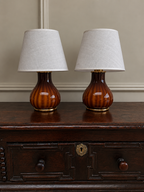 Two matching table lamps with tapered off-white fabric shades and glossy amber-brown ribbed glass bases, displayed on a dark wooden chest against a beige wall.