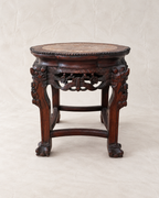 An antique dark brown carved wooden stool with a circular stone or wood top, ornate legs, and claw-foot ends, photographed against a neutral beige backdrop.