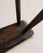 Close-up of a dark stained wooden chair leg joint with a thick rectangular base and two vertical legs, photographed against a light fabric backdrop.