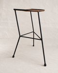 A mid-century modern bar stool with a black metal frame and a woven brown and beige seat, photographed against a neutral off-white fabric backdrop.