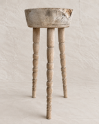 A rustic, distressed wooden stool with a round bowl-like top and three turned spindle legs, shown against a neutral, textured beige backdrop.