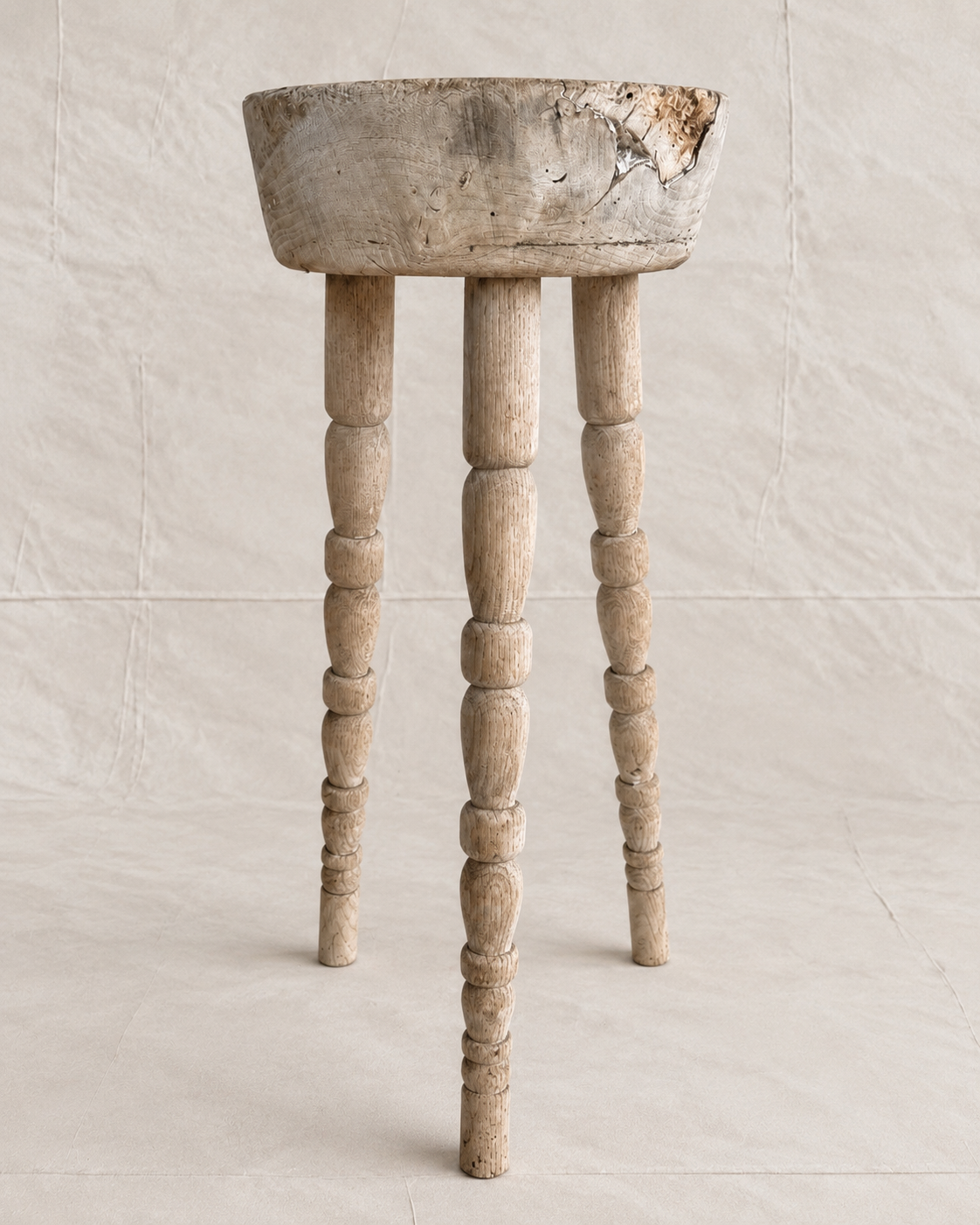 A rustic, distressed wooden stool with a round bowl-like top and three turned spindle legs, shown against a neutral, textured beige backdrop.