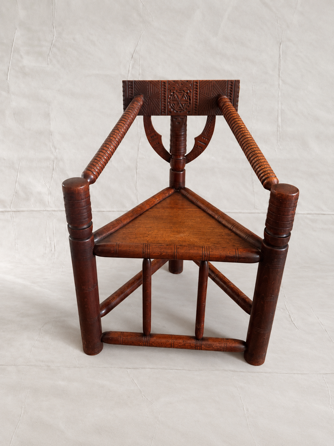A traditional wooden chair with a triangular seat, carved backrest, and woven rope armrests, shown against a light gray fabric backdrop.