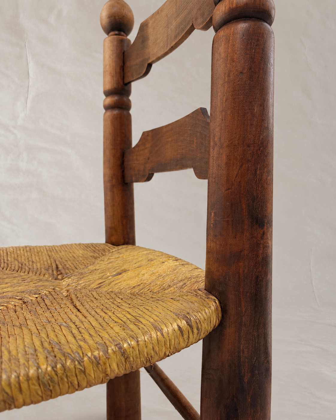 A close-up of a wooden chair showing the side profile, including a dark brown wood frame with a woven yellow-brown seat, set against a light gray fabric backdrop.
